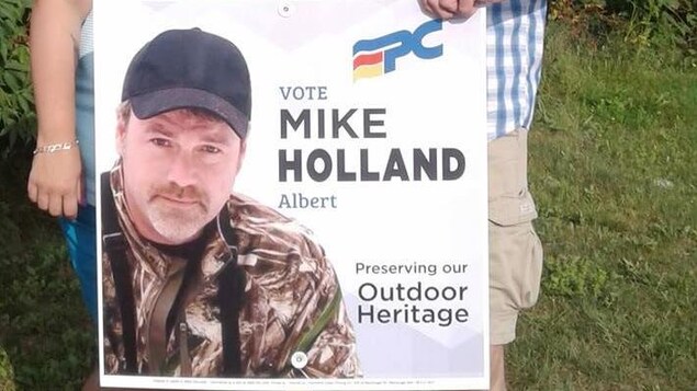Deux personnes hors champs tiennent une affiche électorale de Mike Holland. Deux personnes hors champs tiennent une affiche électorale de Mike Holland.