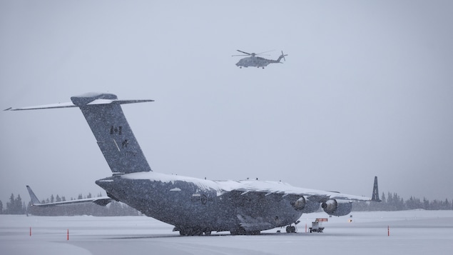 Un avion militaire canadien est à l’arrêt sur une piste enneigée, tandis qu’un hélicoptère militaire vole au-dessus.