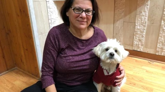 Millie Knapp porte un chandail violet, elle a des lunettes, les cheveux en carré mi-long brun et elle sourit, elle est assise en tailleur et un petit chien blanc qui porte un gilet rouge est à côté d'elle Millie Knapp porte un chandail violet, elle a des lunettes, les cheveux en carré mi-long brun et elle sourit, elle est assise en tailleur et un petit chien blanc qui porte un gilet rouge est à côté d'elle