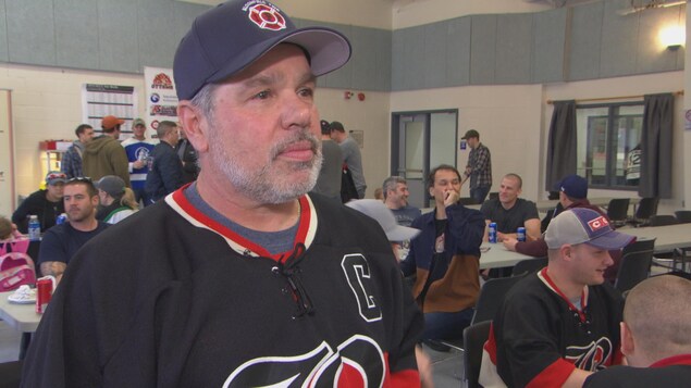 Un homme barbu portant une casquette et un chandail de hockey dans une salle de réception. Un homme barbu portant une casquette et un chandail de hockey dans une salle de réception.