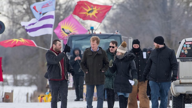 Un groupe de personnes marchent dans la neige près de voies ferrées où des drapeaux autochtones flottent dans le vent. 