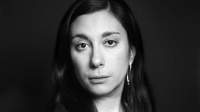 Sur cette photo en noir et blanc, une femme aux longs cheveux foncés et portant un haut noir sans manche regarde la caméra le visage tourné vers la droite.