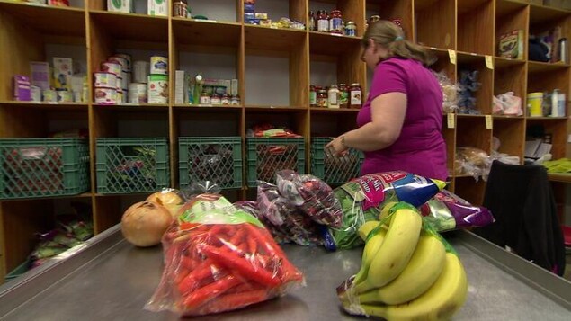 Une femme assise devant des étagères remplies de denrées alimentaires. Au premier plan, on voit des fruits et des légumes frais.