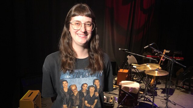 Une percussionniste portant un t-shirt des Backstreet Boys est debout, les mains derrière les dos, à côté d'une batterie. Elle regarde directement la caméra et sourit pour une photo.