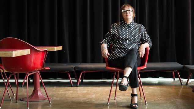 Une photo de Monique Savoie assise sur une chaise rouge d'allure industrielle. Une photo de Monique Savoie assise sur une chaise rouge d'allure industrielle.