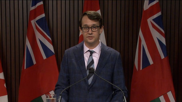 وزير العمل في أونتاريو مونتي ماكنوتون يتحدث في مؤتمر صحفي ونراه واقفاً، أمامه ميكروفونان وخلفه علم كندا وعلمان لأونتاريو.