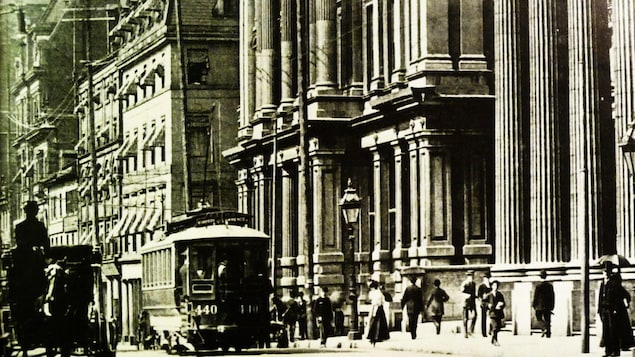 Photo d'archives montrant une rue de Montréal en 1890 avec un tramway, une calèche tirée par un cheval et des passants sur un trottoir devant d'imposantes façades Photo d'archives montrant une rue de Montréal en 1890 avec un tramway, une calèche tirée par un cheval et des passants sur un trottoir devant d'imposantes façades