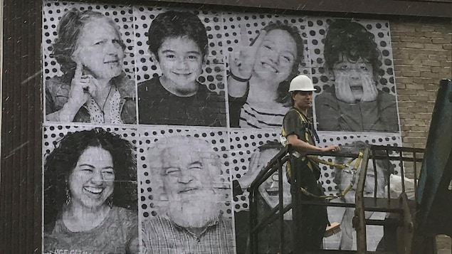 Un homme est en train d'installer une murale sur laquelle on voit des visages souriants de tous âges.