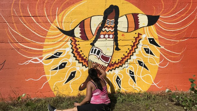 Une artiste peint sur une murale, assise au sol.