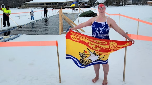 Nadine Bennett en maillot de bain et pieds nus sur la glace pose pour la photo avec un drapeau du Nouveau-Brunswick dans les mains. Derrière elle, un trou a été fait dans la glace pour y faire une ligne de nage. 