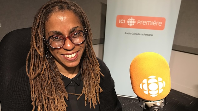 Une femme noire avec des lunettes devant un micro de Radio-Canada sourit dans un studio.