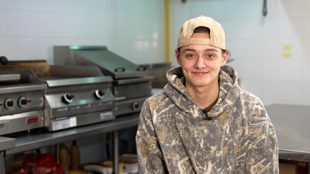 Un jeune homme souriant, portant une casquette beige à l'envers et un coton ouaté à motif de camouflage, pose devant des équipements de cuisine professionnelle en acier inoxydable, incluant des plaques de cuisson et des friteuses, sous un éclairage néon.
