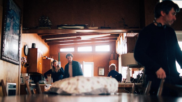 Un homme, une femme et un bébé sont dans la cuisine, alors qu'un autre homme en avant-plan est proche de la table à manger. 