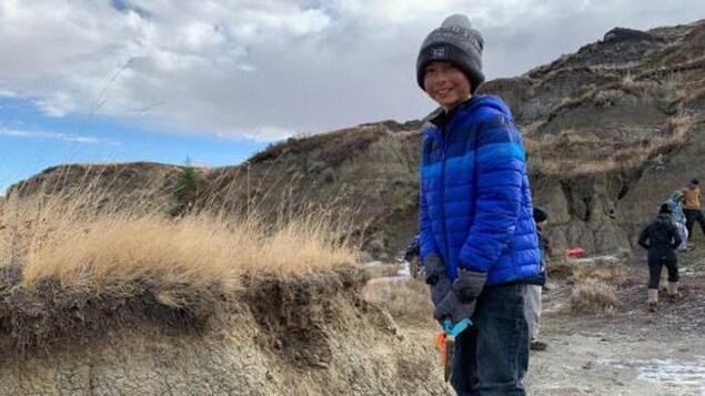 Un jeune enfant, tuque, gant et veste, tient une petite pelle devant le site d'excavation, typique des Badlands albertaines.