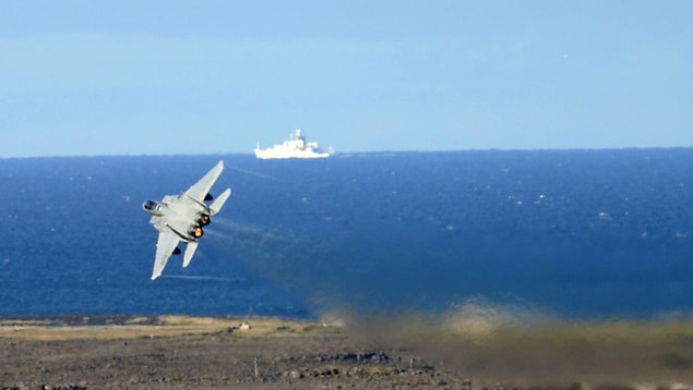 A fighter jet  flying with a boat on the water in the background.