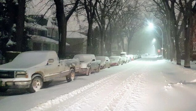 Des voitures stationnées dans une rue enneigée en soirée.