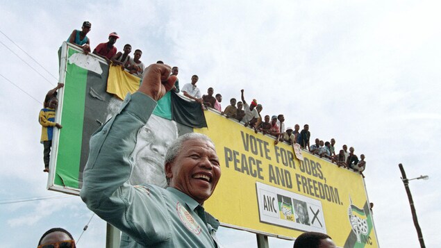 Souriant et le poing levé, Nelson Mandela se tient devant un panneau sur lequel se tiennent de nombreuses personnes.
