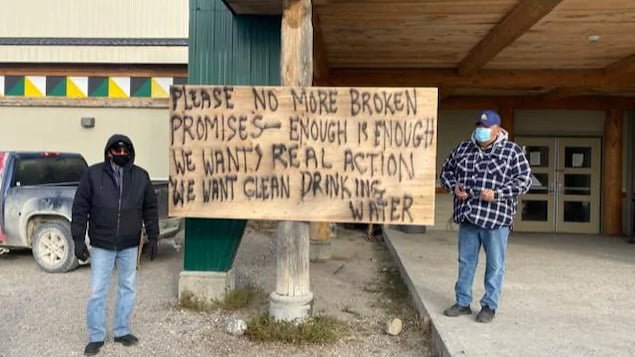 Deux hommes de chaque côté d'une pancarte sur laquelle est écrit « S'il vous plaît, plus de promesses non tenues, assez c'est assez ».