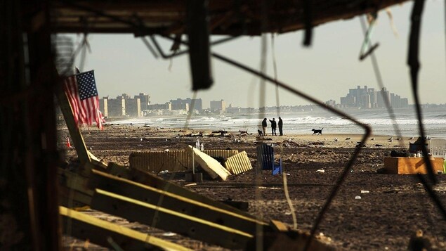 Des infrastructures brisées et un drapeau américain déchiré se trouvent le long d'une plage jonchées de déchets. Des infrastructures brisées et un drapeau américain déchiré se trouvent le long d'une plage jonchées de déchets.