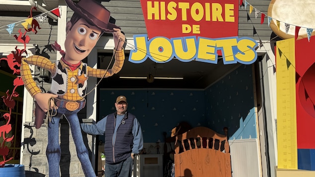 Un homme se tient debout devant sa maison, à côté d'un personnage de Woody géant. Il y a une affiche inscrite Histoire de jouets.