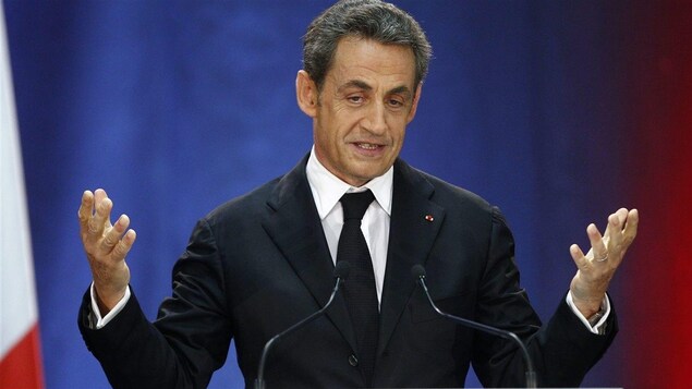 Nicolas Sarkozy debout derrière un pupitre et des micros, les avant-bras levés.