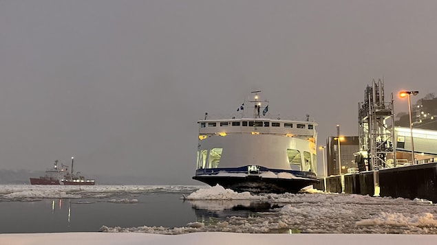 Un traversier accosté à un quai du côté de Québec, en hiver. À l’arrière-plan, un navire de la Garde côtière navigue sur le fleuve Saint-Laurent.