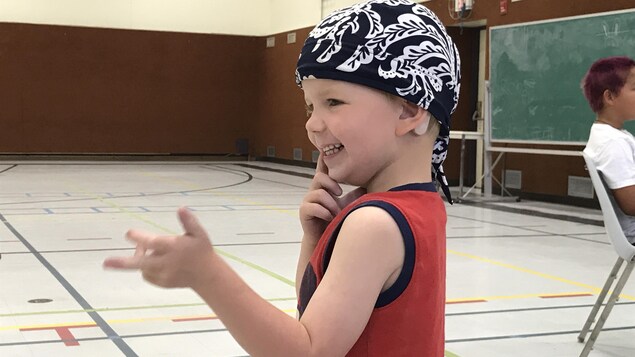 Le petit garçon Noah de trois ans porte un foulard sur la tête et sourit avec un doigt à la bouche.