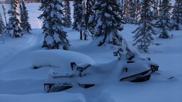 une motoneige en hiver, entourée de neige. Derrière, une ligne de conifères, au loin un lac gelé et ennneigé et la ligne d'horizon. Devant, des taces dans la neige qui foirme un sentier menant vers la moto. La moto est elle-même sous de la neige qui n'a pas encore été déblayée. une motoneige en hiver, entourée de neige. Derrière, une ligne de conifères, au loin un lac gelé et ennneigé et la ligne d'horizon. Devant, des taces dans la neige qui foirme un sentier menant vers la moto. La moto est elle-même sous de la neige qui n'a pas encore été déblayée.