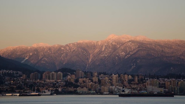 D'imposantes montagnes enneigées avec un lever de soleil et une ville avec des gratte-ciel devant une étendue d'eau.
