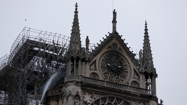 Des pompiers lancent de un immense jet d'eau vers le toit de la cathédrale. Le haut du mur du devant est noirci par le passage des flammes.