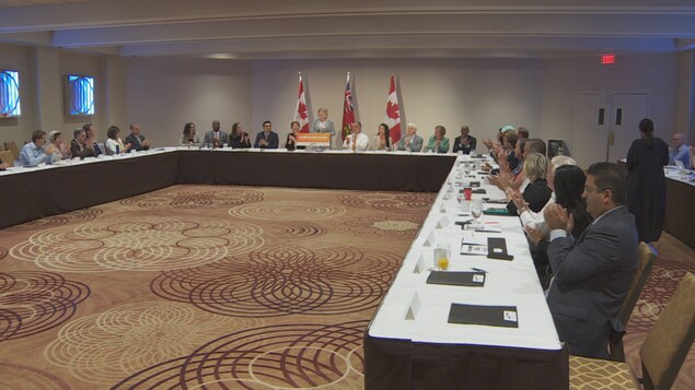 Andrea Horwath debout, avec autour d'elle ses députés assis autour de tables. Andrea Horwath debout, avec autour d'elle ses députés assis autour de tables.