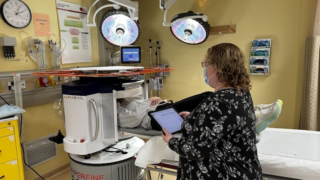 Une femme portant un masque médical tient une tablette numérique dans ses mains pendant qu'une patiente est couchée sur le dos lors d'un examen par imagerie par résonance magnétique.
