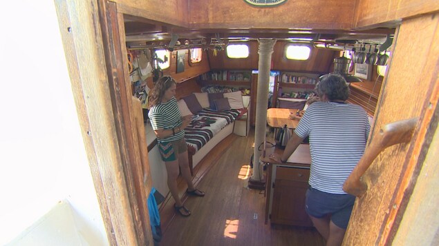 Le bateau qui contient deux chambres, une salle de bain, une cuisine et quelques espaces de vie commune. La photo est prise de l'extérieur, vers l'intérieur du bateau. On voit une cinquantaine de livres dans une bibliothèque, un divan avec de nombreux coussins. Cheryl lit quelque chose sur son téléphone. Le bateau qui contient deux chambres, une salle de bain, une cuisine et quelques espaces de vie commune. La photo est prise de l'extérieur, vers l'intérieur du bateau. On voit une cinquantaine de livres dans une bibliothèque, un divan avec de nombreux coussins. Cheryl lit quelque chose sur son téléphone.