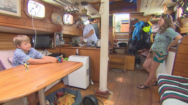 Moment en famille chez les Nuttall-Sloane. Gordon prépare un café pendant que Cheryl discute avec leur fils, Strummer, qui joue à des jeux sur une table au centre du bateau. On voit une cuisinette et un divan. Des objets sont accrochés au plafond et posés sur le bord des hublots. Moment en famille chez les Nuttall-Sloane. Gordon prépare un café pendant que Cheryl discute avec leur fils, Strummer, qui joue à des jeux sur une table au centre du bateau. On voit une cuisinette et un divan. Des objets sont accrochés au plafond et posés sur le bord des hublots.