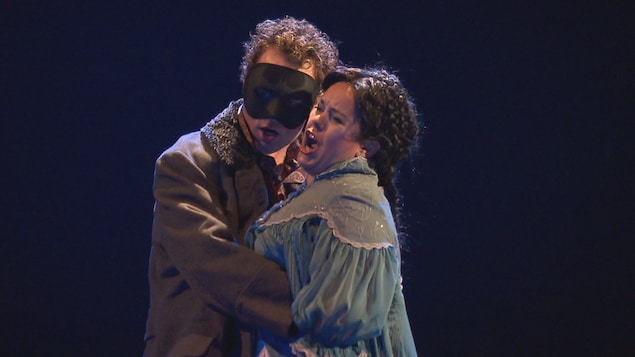 Deux chanteurs d'opéra chantent, dans les bras l'un de l'autre. L'homme a un masque sur les yeux et la femme a les cheveux tressés. 