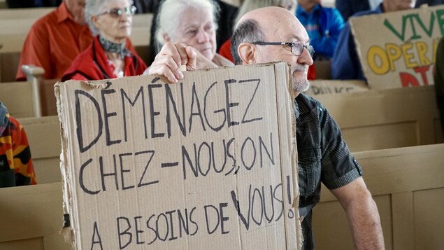 Un homme tient une pancarte où on peut lire : Déménagez chez-nous, on a besoin de vous!          