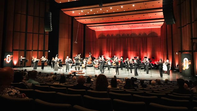 Des musiciens debout sur une scène, devant un public, dans une salle aux teintes rouge