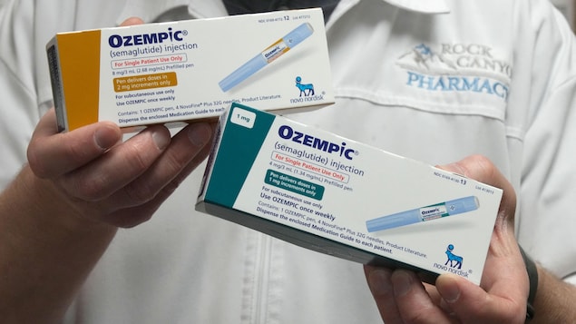 Un homme vêtu d'un sarrau de pharmacien tient dans ses mains deux boîtes d'Ozempic, qui affichent des informations sur le médicament et une photo d'un injecteur.