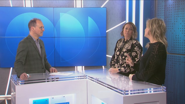 Trois personnes sont dans un studio de télévision moderne. À gauche, un homme en veston discute avec deux femmes debout derrière un comptoir blanc en forme de L. L'arrière-plan présente des écrans bleus avec des motifs circulaires.