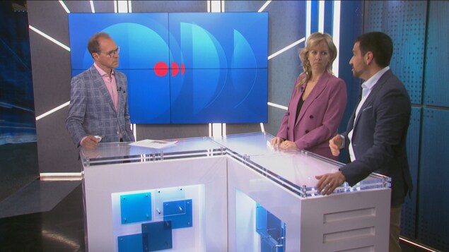 Trois personnes, deux hommes et une femme, discutent sur un plateau de télévision moderne. Ils se tiennent derrière un pupitre blanc en forme de L, devant un grand écran bleu affichant des graphiques abstraits.