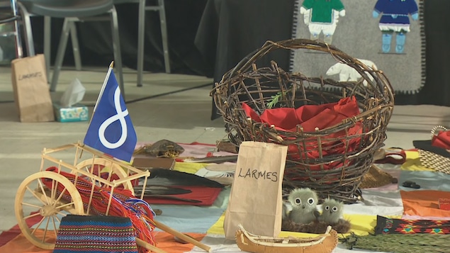 Un panier, un canot miniature en écorce, de petits jouets et un sac brun où il est écrit « Larmes ». Un panier, un canot miniature en écorce, de petits jouets et un sac brun où il est écrit « Larmes ».