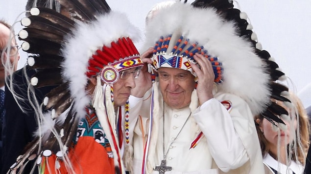 البابا فرنسيس (إلى اليمين) يعتمر غطاء رأس تقليدياً للسكان الأصليين تلقاه في نهاية خطابه في ماسكواسيس في ألبرتا في 25 تكموز (يوليو) 2022 في إطار زيارته إلى كندا.