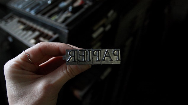 Une main tient une série de caractères typographiques en métal destinés à imprimer le mot «papier».