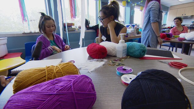 Une fillette et une jeune femme sont assises à une table, sur laquelle sont posées des pelotes de laine, des ciseaux et d'autres accessoires.