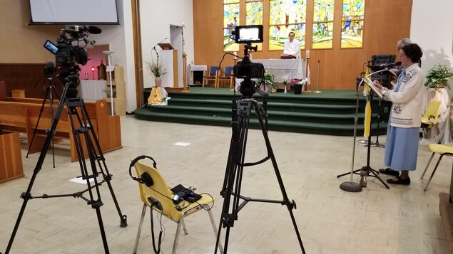 Dans une église, des caméras filment un abbé et des chanteurs, tous à bonne distance les uns des autres.