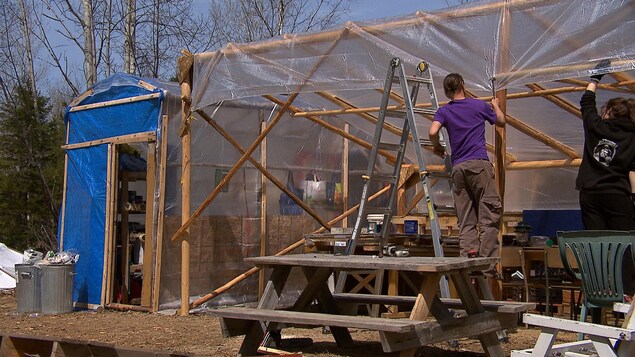 Des occupants du campement près d'un chemin d'accès au site Galt de la pétrolière Junex travaillent sur leurs installations. Un homme avec un marteau clou un pellicule imperméable sur une structure de bois. Des occupants du campement près d'un chemin d'accès au site Galt de la pétrolière Junex travaillent sur leurs installations. Un homme avec un marteau clou un pellicule imperméable sur une structure de bois.