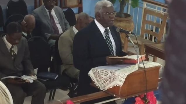 On voit, en plongée, le pasteur haïtiens parler debout devant les fidèles réunis dans l'église. Derrière lui, des hommes sont assis et écoutent ses paroles en lisant les écritures. On voit, en plongée, le pasteur haïtiens parler debout devant les fidèles réunis dans l'église. Derrière lui, des hommes sont assis et écoutent ses paroles en lisant les écritures.