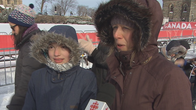 À l'extérieur, une jeune fille et une femme adulte vêtues de manteaux d'hiver répondent aux questions d'un journaliste. Derrière elles, des personnes mettent leurs patins à glace 
