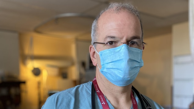 Le docteur Bellemare porte un sarrau bleu, un masque et a un stéthoscope autour du cou.