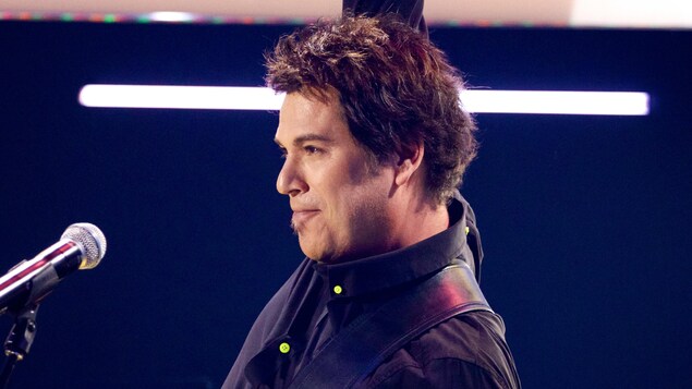 Le chanteur Patrick Bourgeois, cheveux courts, guitare à la main en performance à l'émission « C'est ma toune » en 2014. Un micro se tient devant lui. Le chanteur Patrick Bourgeois, cheveux courts, guitare à la main en performance à l'émission « C'est ma toune » en 2014. Un micro se tient devant lui.